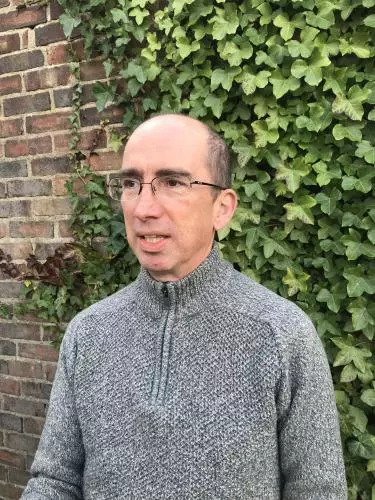 Photo of Neil Kenny standing in front of a brick wall covered in ivy. He is looking off camera to the left.