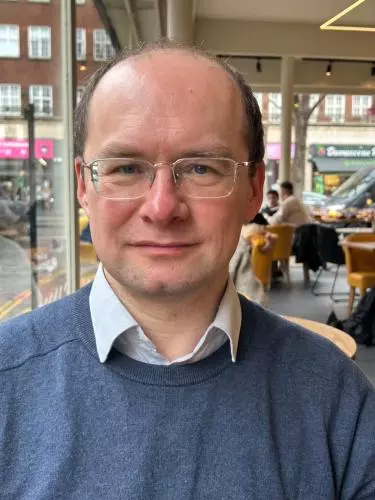 Portrait of Andrii Pastushenko wearing glasses, a blue sweater, and a collared shirt, seated indoors in a café