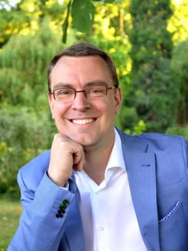 Professor Wim Decock smiling to camera, wearing glasses, a white shirt, and a light blue blazer, resting his chin on his hand while seated outdoors against a softly blurred green garden background.