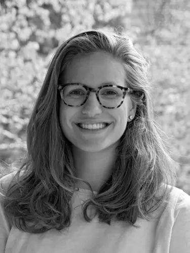 Black-and-white headshot of Hannah Kaemmer with long hair and patterned glasses, wearing a light-colored top, photographed outdoors against a softly blurred leafy background.