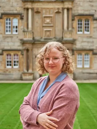 Kathryn Hempstead standing in Fellows' Quad, Merton College. Smiling to camera with arms crossed, wearing a pink cardigan and a blue shirt dress.