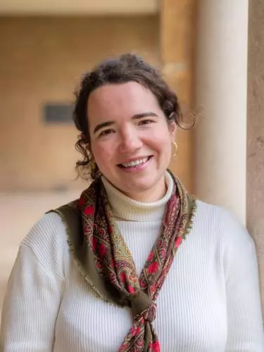 Portrait of Marie Raulier, smiling, wearing a white turtleneck and patterned scarf, standing beside a large stone column.