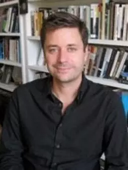 Photo of Adam Smyth smiling to camera with a bookcase behind him