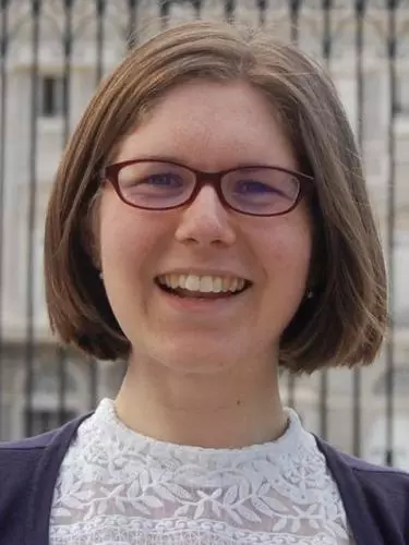 Photo of Alice Brooke smiling to camera. Metal bars and a stone building can be seen behind her. 