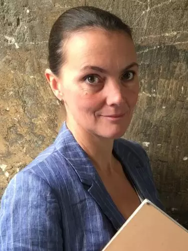 Photo of Suzanne Aspden looking to camera holding a book or score, standing in front of a stone wall
