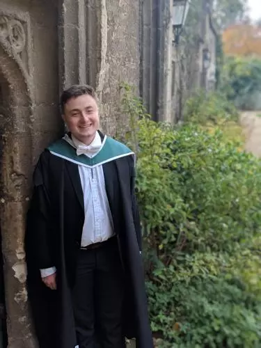 Photo of Ben Wilkinson Turnbull smiling to camera, standing in front of a stone archway. Foliage and a stone wall is visible through the archway.