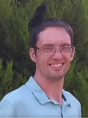 Photo of Dr Johnathan Patterson smiling to camera, standing outside in front of foliage. 