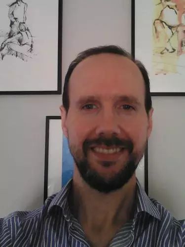 Photo of Gonzalo Rodriguez-Pereyra smiling to camera. Framed paintings are visible on the white wall behind him.