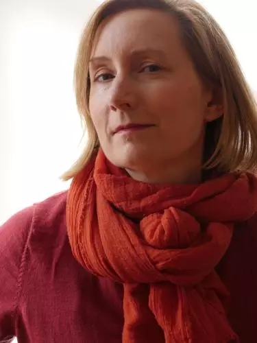Katherine Ibbett looking to camera, sitting in front of a white wall with the reflection of a window behind her. 