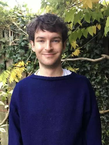 Photo of Alex Laar smiling to camera. He is standing outside, in front of an ivy covered fence. Foliage and a tree branch is visible behind.