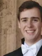 Photo of Samuel Teague smiling to camera in front of a stone wall and a window.