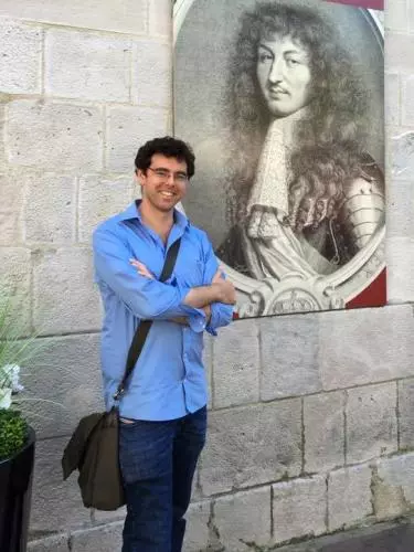 Photo of Jonathan Healey smiling, leaning against a wall with a large reproduction of an early modern engraved portrait. 