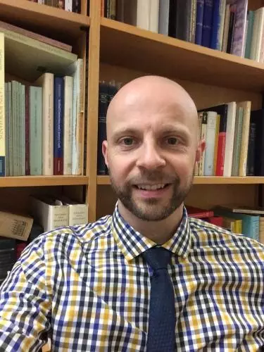 Photo of Daniel Wakelin smiling to camera with a bookcase behind him