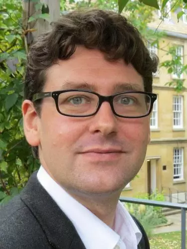 Photo of George Southcombe looking to camera, standing outside with foliage, a lawn and a stone building visible behind him.
