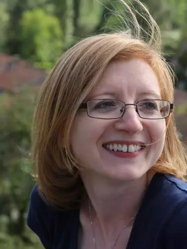 Photo of Helen Swift smiling, looking to the right of the camera. She is outside, with trees and houses visible in the background. 