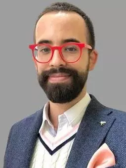 Photo of Dr Vincent Roy Di Piazza. Wearing blue jacket, cream vest and red glasses. Bearded. Looking directly at camera.
