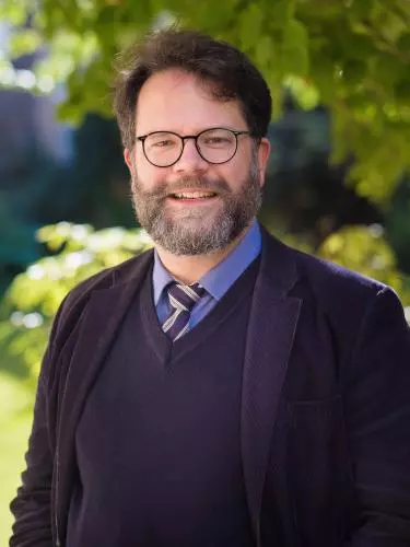 Photo of Dr Christoph Schmitt-Maass wearing a dark suit with blue and yellow tie. Bearded and wearing glasses, smiling at camera.