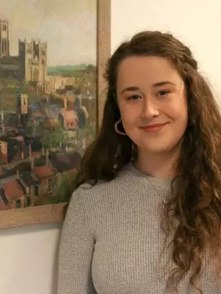 Photo of Clare Burgess smiling to camera, standing in front of a painting of a city. 