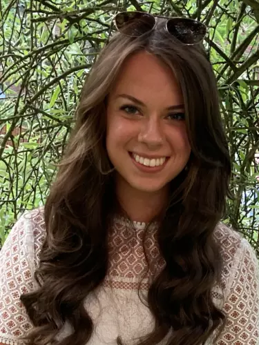 Photo of Caroline Taylor. Smiling directly at the camera, wearing white dress and sunglasses atop head.