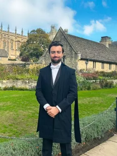 Photo of Samuel Head in dark navy Oxford subfusc. Standing before a green and limestone buildings, likely Christ Church.