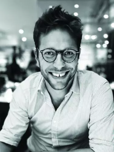 Black and white photo of Olly Markeson. Wearing white shirt and glasses. Blurred restaurant background.