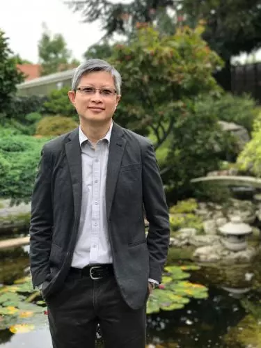 Photo of Tian Yuan Tan standing in front of a pond of lilypads. A bridge and trees are visible in the background. 