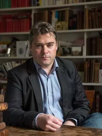 Photo of Will Poole sitting at a desk. There is a pile of early modern books on the desk in front of him, and bookshelves behind him. 