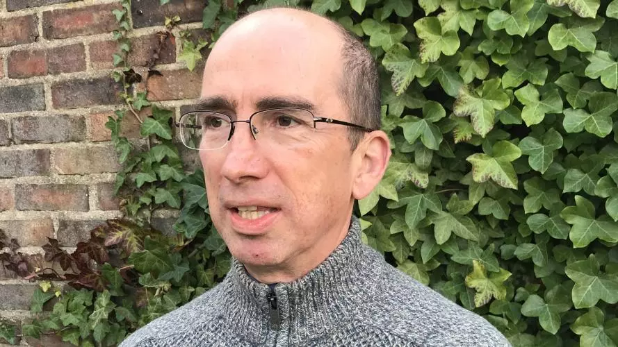 Photo of Neil Kenny standing in front of a brick wall covered in ivy. He is looking off camera to the left.