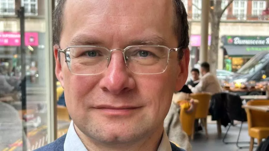 Portrait of Andrii Pastushenko wearing glasses, a blue sweater, and a collared shirt, seated indoors in a café