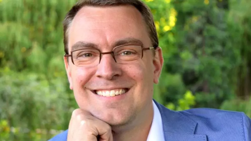 Professor Wim Decock smiling to camera, wearing glasses, a white shirt, and a light blue blazer, resting his chin on his hand while seated outdoors against a softly blurred green garden background.