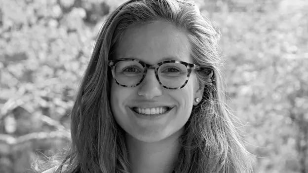 Black-and-white headshot of Hannah Kaemmer with long hair and patterned glasses, wearing a light-colored top, photographed outdoors against a softly blurred leafy background.