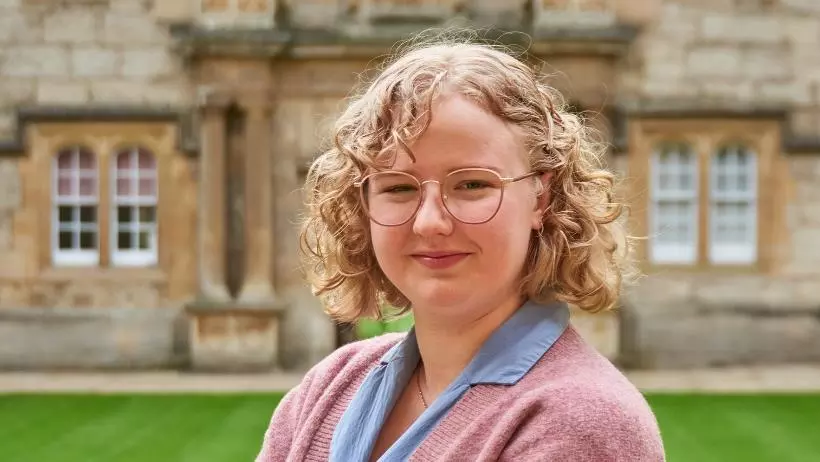 Kathryn Hempstead standing in Fellows' Quad, Merton College. Smiling to camera with arms crossed, wearing a pink cardigan and a blue shirt dress.