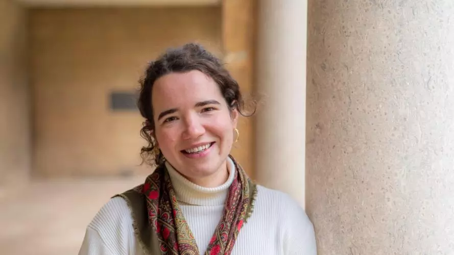 Portrait of Marie Raulier, smiling, wearing a white turtleneck and patterned scarf, standing beside a large stone column.