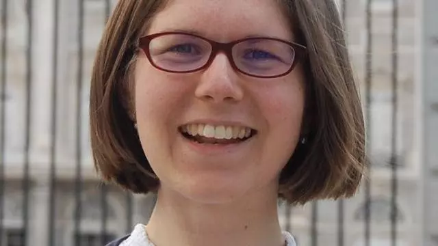 Photo of Alice Brooke smiling to camera. Metal bars and a stone building can be seen behind her. 