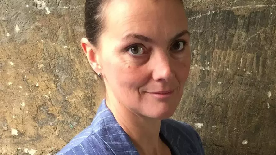 Photo of Suzanne Aspden looking to camera holding a book or score, standing in front of a stone wall