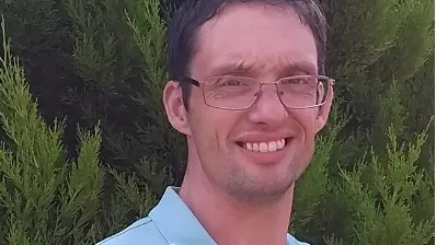 Photo of Dr Johnathan Patterson smiling to camera, standing outside in front of foliage. 