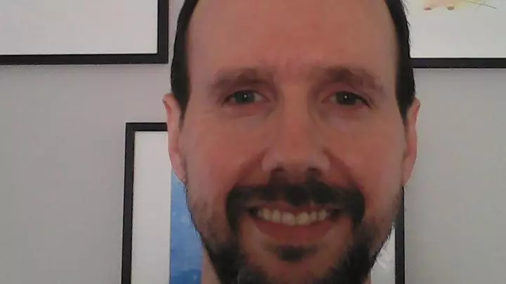 Photo of Gonzalo Rodriguez-Pereyra smiling to camera. Framed paintings are visible on the white wall behind him.