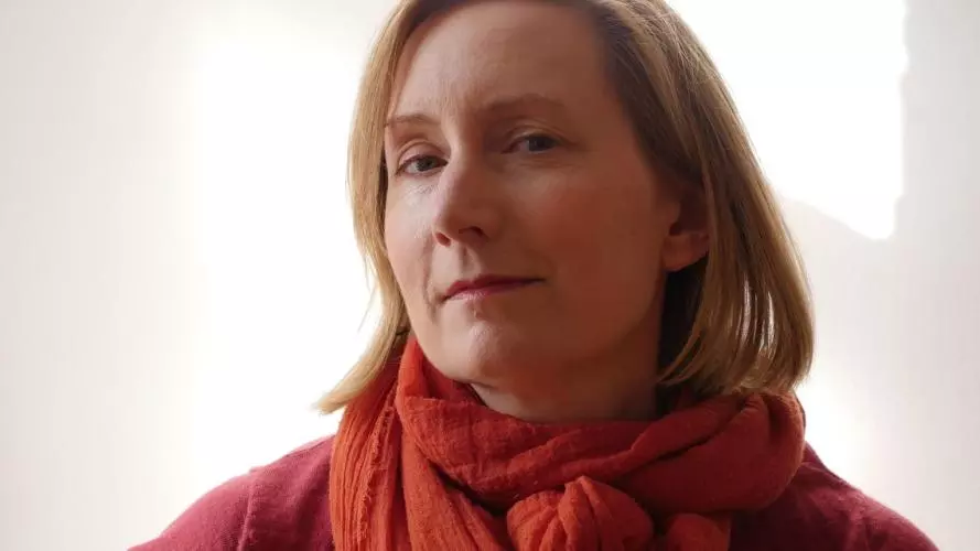 Katherine Ibbett looking to camera, sitting in front of a white wall with the reflection of a window behind her. 