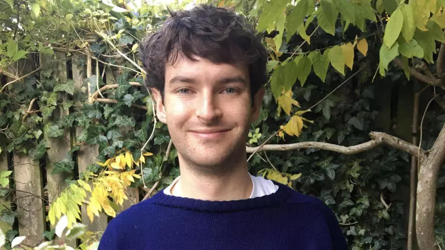 Photo of Alex Laar smiling to camera. He is standing outside, in front of an ivy covered fence. Foliage and a tree branch is visible behind.