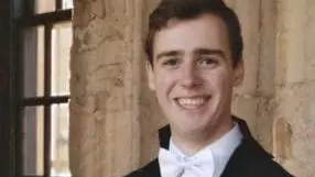 Photo of Samuel Teague smiling to camera in front of a stone wall and a window.