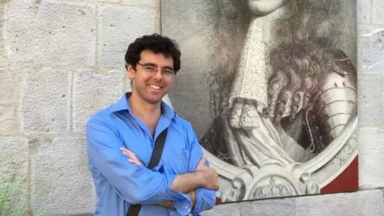 Photo of Jonathan Healey smiling, leaning against a wall with a large reproduction of an early modern engraved portrait. 