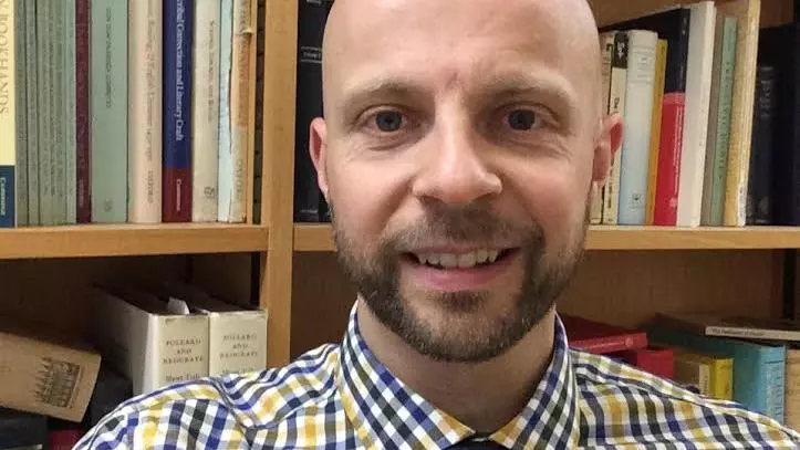 Photo of Daniel Wakelin smiling to camera with a bookcase behind him
