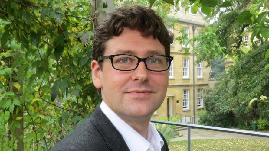 Photo of George Southcombe looking to camera, standing outside with foliage, a lawn and a stone building visible behind him.