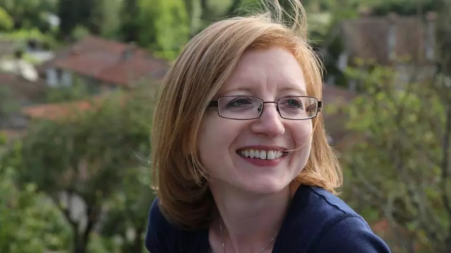 Photo of Helen Swift smiling, looking to the right of the camera. She is outside, with trees and houses visible in the background. 