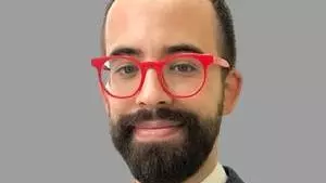 Photo of Dr Vincent Roy Di Piazza. Wearing blue jacket, cream vest and red glasses. Bearded. Looking directly at camera.