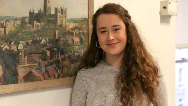 Photo of Clare Burgess smiling to camera, standing in front of a painting of a city. 
