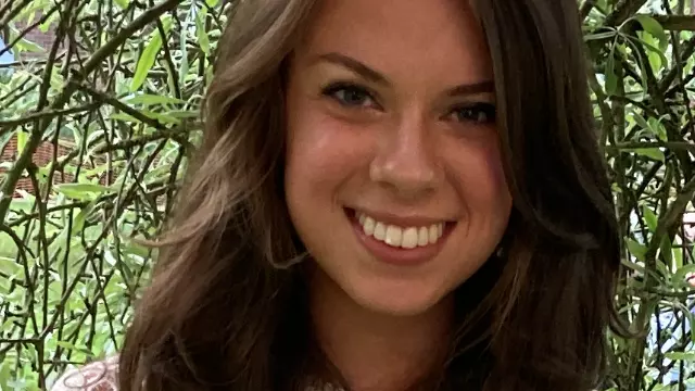 Photo of Caroline Taylor. Smiling directly at the camera, wearing white dress and sunglasses atop head.
