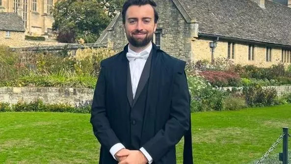 Photo of Samuel Head in dark navy Oxford subfusc. Standing before a green and limestone buildings, likely Christ Church.