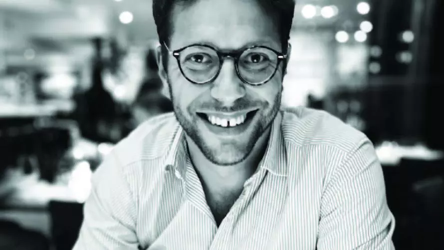 Black and white photo of Olly Markeson. Wearing white shirt and glasses. Blurred restaurant background.