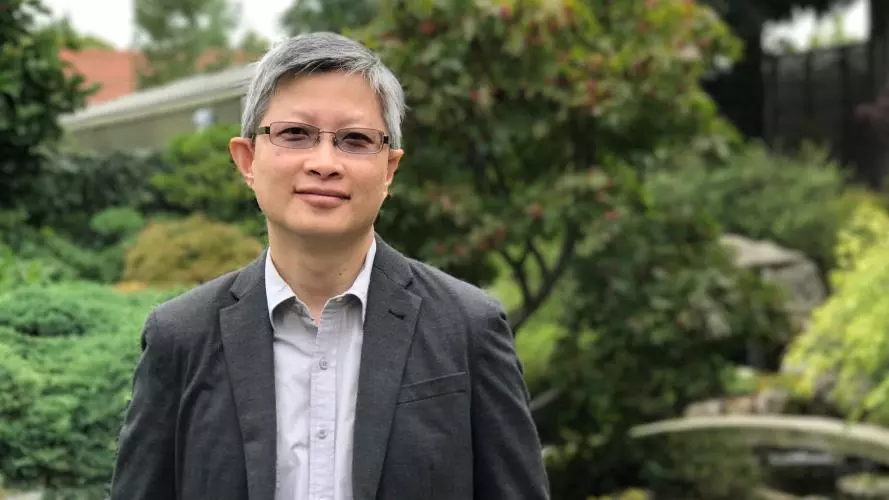 Photo of Tian Yuan Tan standing in front of a pond of lilypads. A bridge and trees are visible in the background. 
